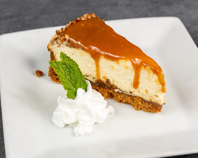 A slice of cheesecake with caramel drizzle, whipped cream, and a mint leaf on a white plate.