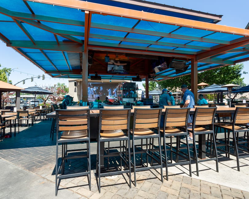 Outdoor bar area with high-top tables and stools under a blue canopy, with patrons visible.