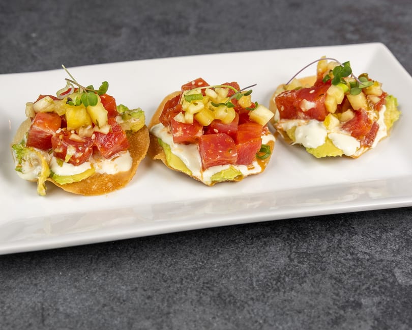 Three tuna poke tostadas with avocado, pineapple, and microgreens on a white rectangular plate.