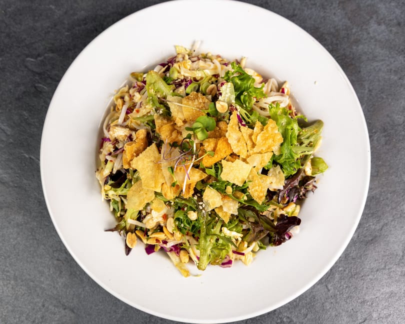 Asian chicken salad with mixed greens, noodles, peanuts, and crispy wonton strips in a white bowl.
