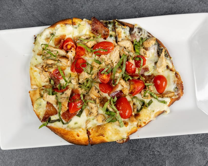 Chicken flatbread with melted cheese, cherry tomatoes, and basil on a white rectangular plate.