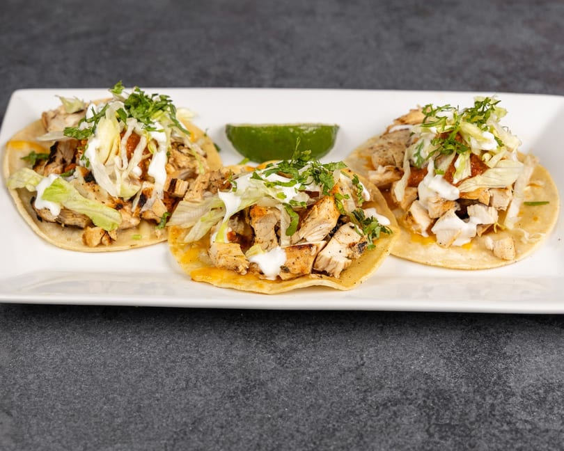 Three chicken tacos with lettuce, sour cream, and cilantro, served with a lime wedge.