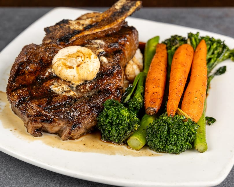 Grilled steak with a pat of butter, served with roasted carrots and broccolini on a white plate.