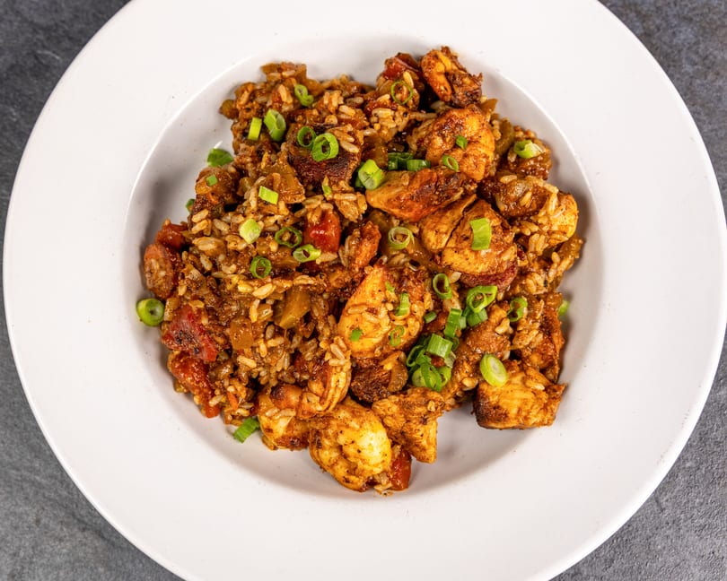 A white bowl filled with jambalaya, featuring rice, chicken, shrimp, and sausage, garnished with green onions.