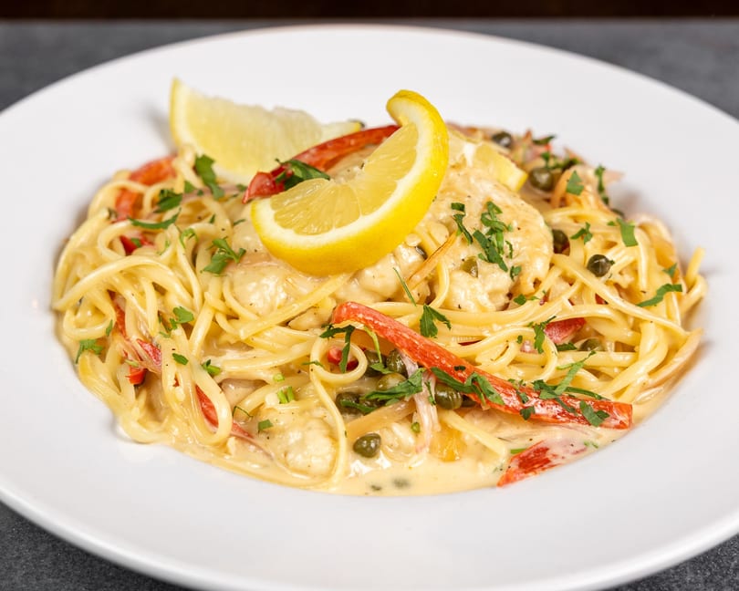 Creamy pasta dish with chicken, red peppers, capers, and parsley, garnished with lemon wedges.