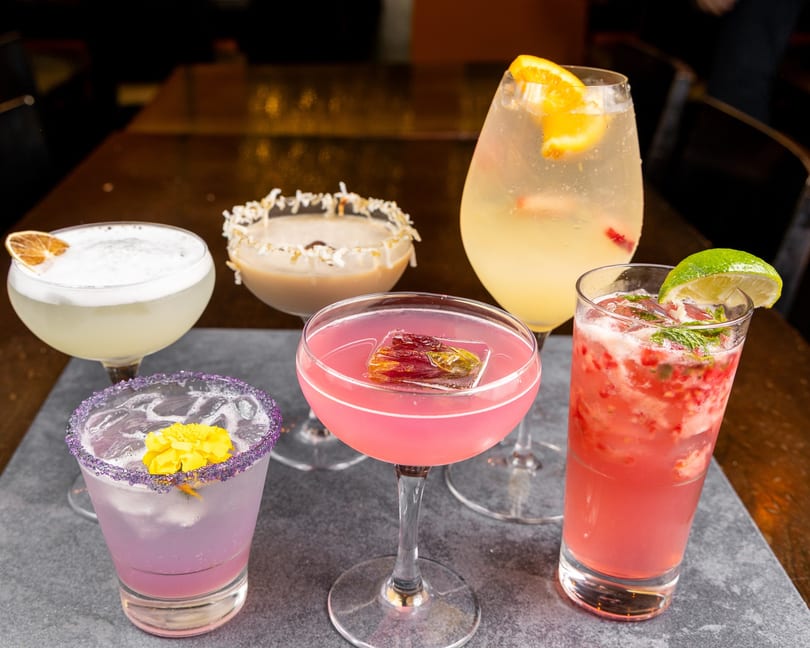 Five colorful cocktails, garnished with fruit and flowers, arranged on a dark stone surface.