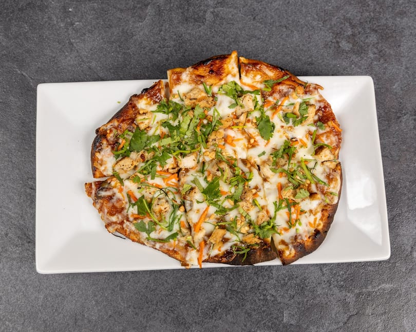 Flatbread pizza with chicken, cheese, carrots, and cilantro, sliced on a white rectangular plate.