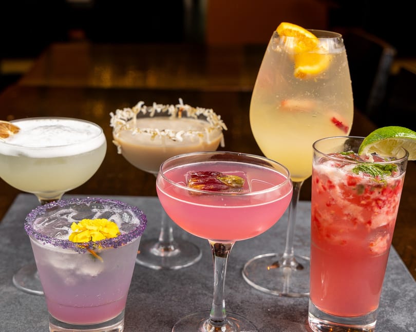 Five colorful cocktails, including pink, purple, and yellow drinks, garnished with fruit and flowers.